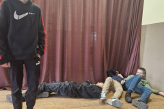 Skečs - zēns stāv uz skatuves pārējie guļ uz zemes